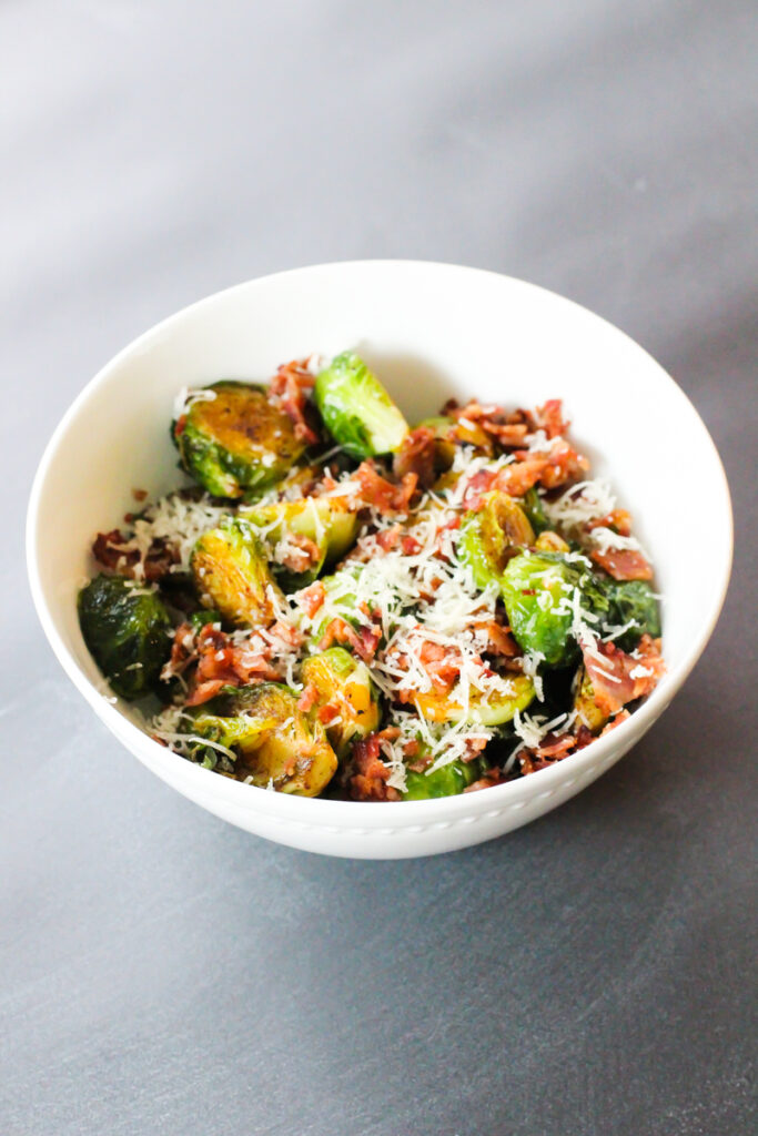 A white china bowl filled with fried Brussels sprouts. The sprouts are topped with crumbled bacon bits and freshly grated parmesan cheese. Background is a gray countertop.