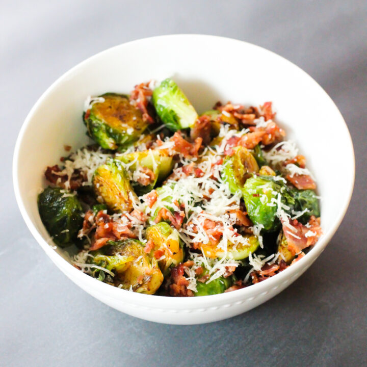 A white china bowl filled with fried Brussels sprouts. The sprouts are topped with crumbled bacon bits and freshly grated parmesan cheese. Background is a gray countertop.